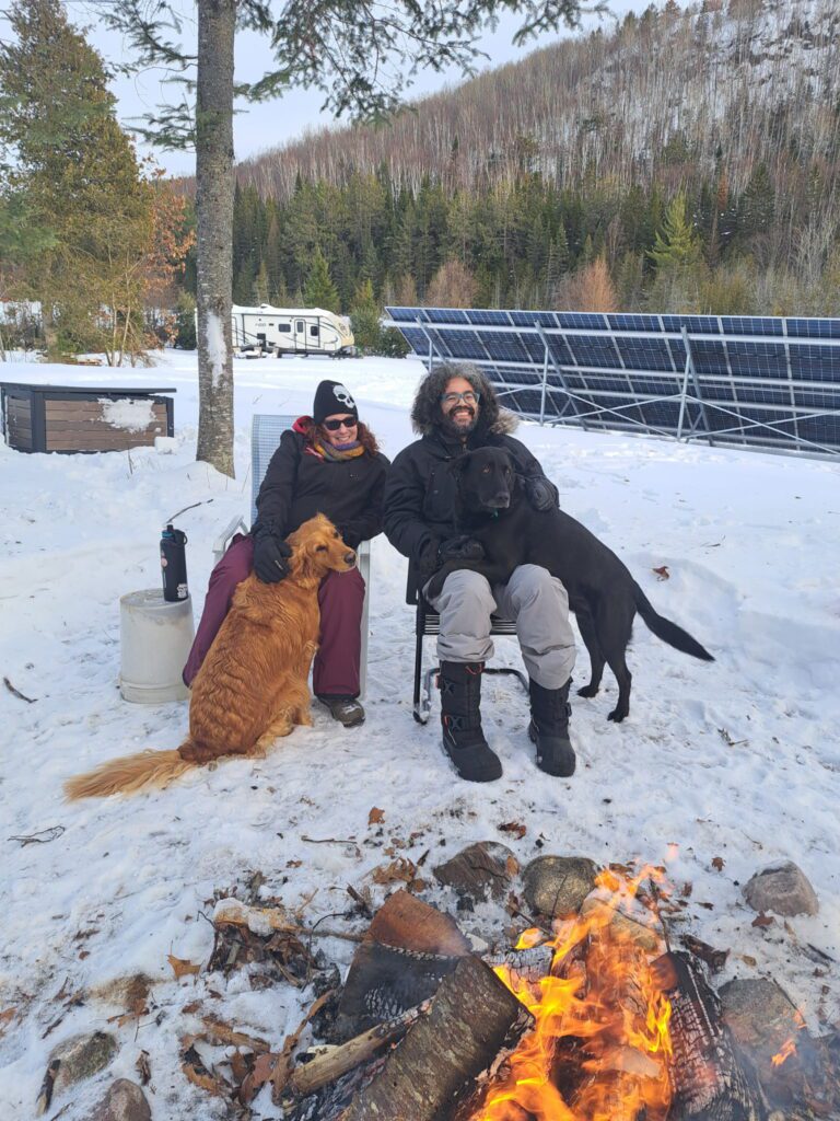 Two people sitting by a campfire in the snow with two dogs.